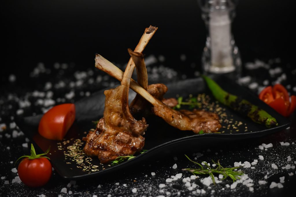 Deliciously presented grilled lamb chops with herbs, tomatoes, and peppers on a black dish.