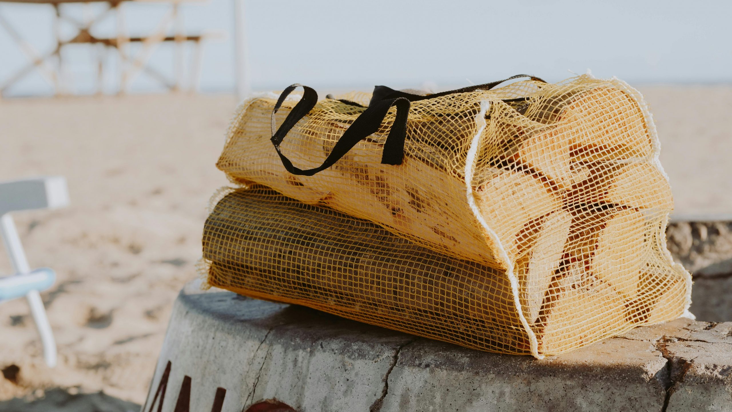 Net bag of firewood resting on a fire pit at the beach. Perfect for a summer outing or bonfire vibe.
